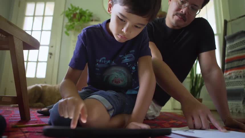 A man and a child work together using a piece of paper and a tablet while sitting on the floor in a room