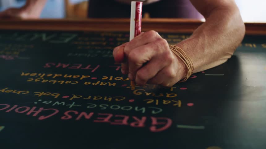 A person writes on a menu board using a yellow chalk marker