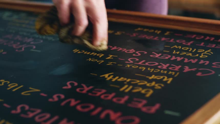 A woman holding a cloth is wiping a blackboard that has food listed on it in purple and yellow
