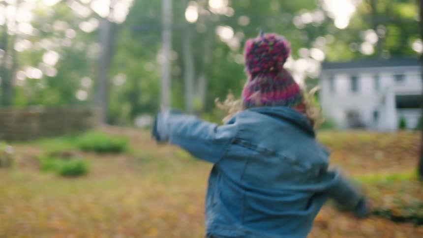 Little girl runs and jumps in pile of leaves on a sunny day with trees and houses in the background