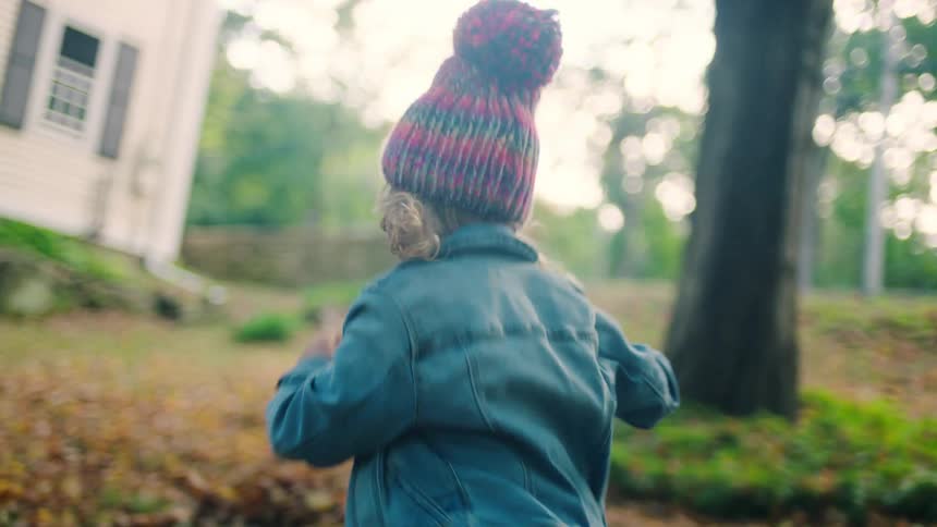 A young girl runs in her backyard full of leaves wearing a jacket and hat
