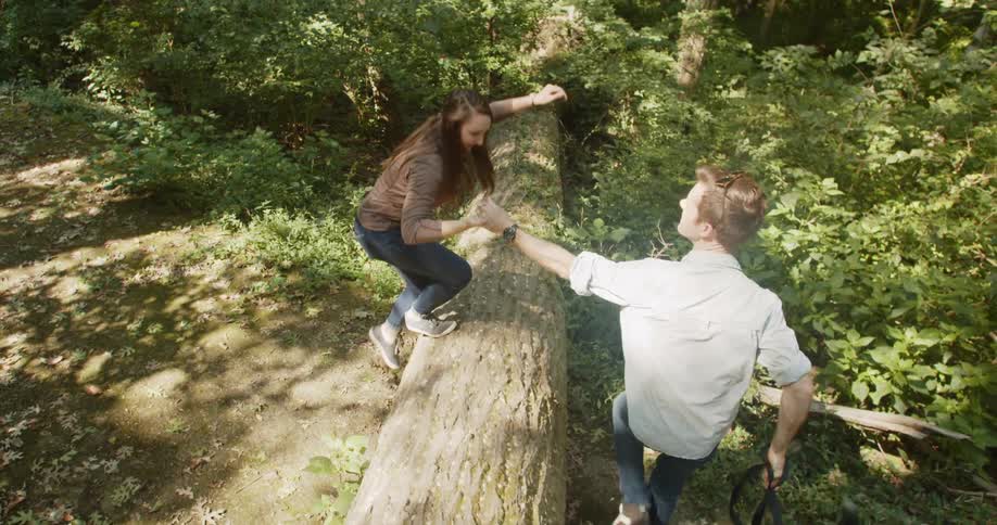 Man holding the hand of a woman helps her walk over a tree trunk in the forest