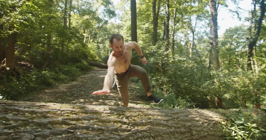Shirtless man with pants and running shoes runs and jumps over a tree trunk in the woods