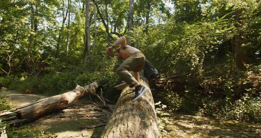 A man not wearing a shirt runs and jumps over a large tree fallen over in the woods