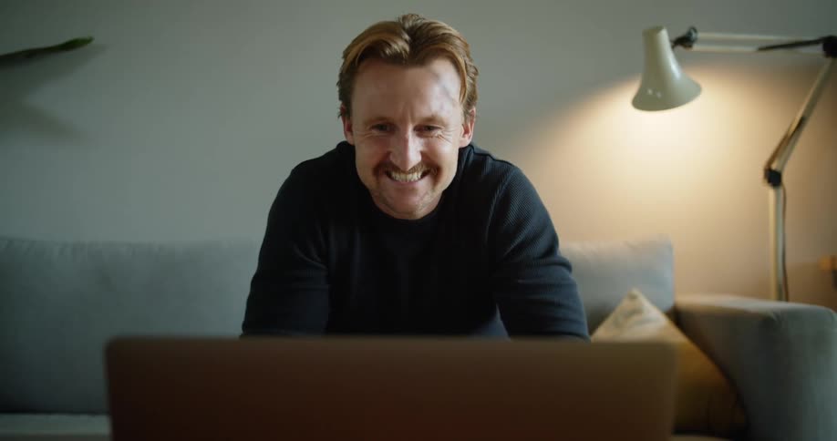 Man sitting in a living room on a sofa video chatting on a laptop computer