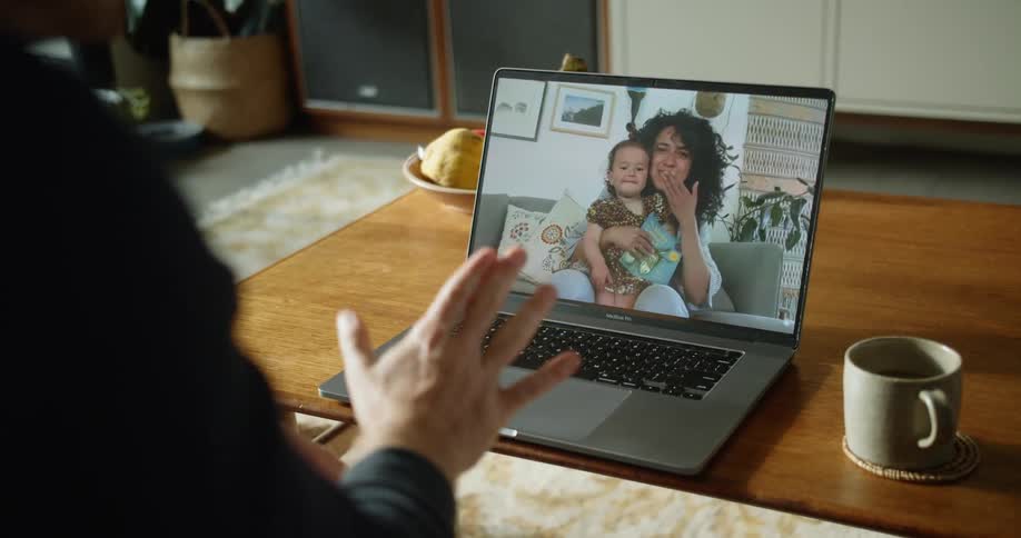 A woman holds a child on the screen of a laptop sitting on a table in front of a man in a room as they throw kisses to one another during the day