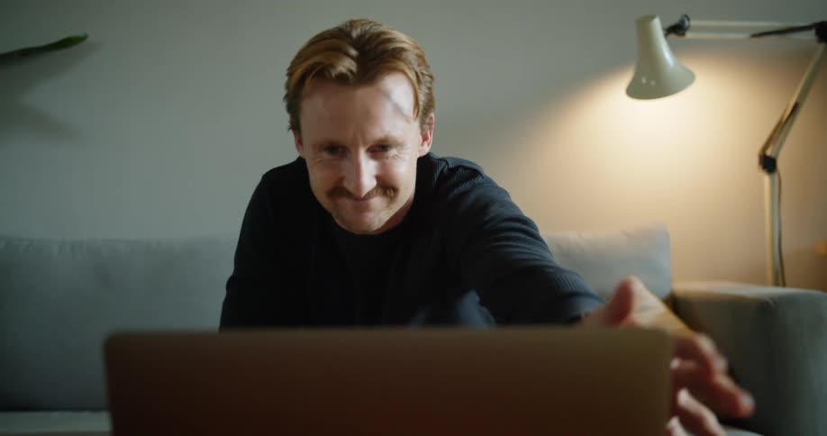 Man sitting in a living on a sofa waving goodbye at a laptop screen then closes it and picks up coffee mug
