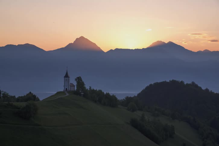 Sun rises over the mountains behind a hillside with a building