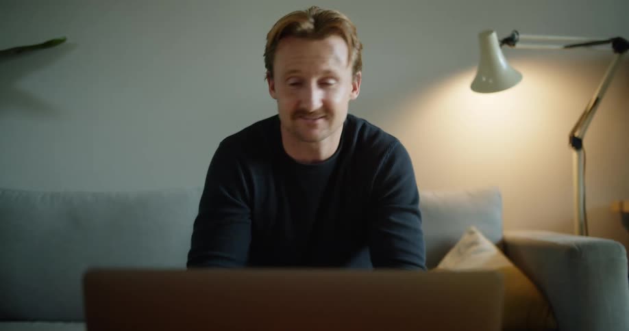 A man smiles as he sits on his couch and talks to someone on a video call on his laptop