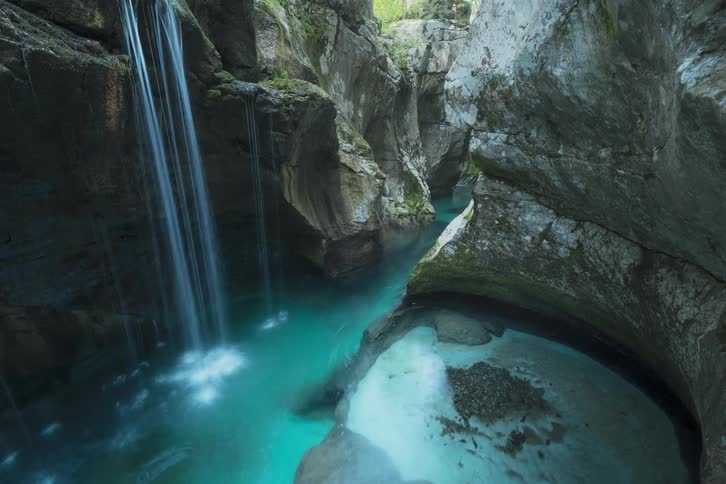 Waterfall flows into grotto joining water flowing through break in cave wall showing vegetation and sunshine