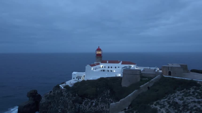 People wander past parked car with its headlights on near building standing on promontory with attached lighthouse