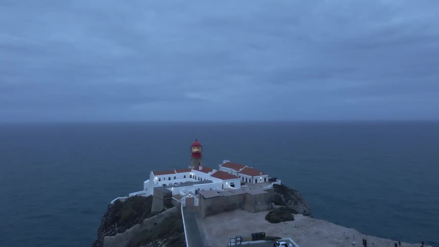 Aerial view of a lighthouse building on the edge of cliff next to the ocean with people walking down below