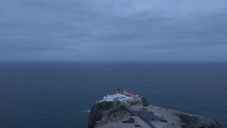 People walk around near parking lot of building complex with attached lighthouse sitting on promontory over ocean