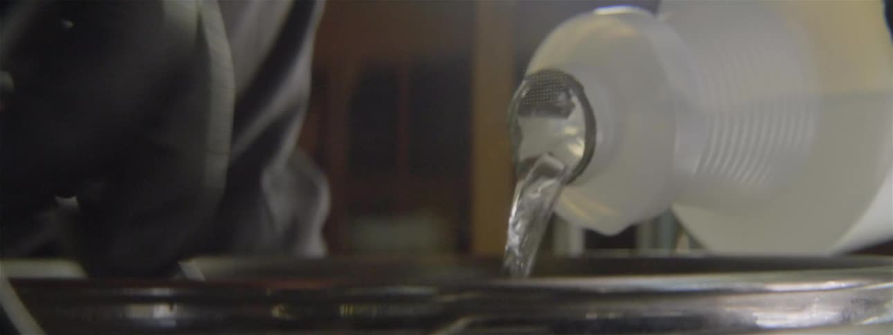 Hand pouring a clear solution into a metal pot and stirring a spoon with the other hand