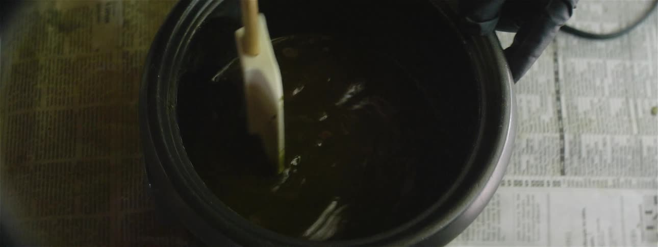 Hands wearing black gloves stirring a green liquid in a pot with a mixing spatula