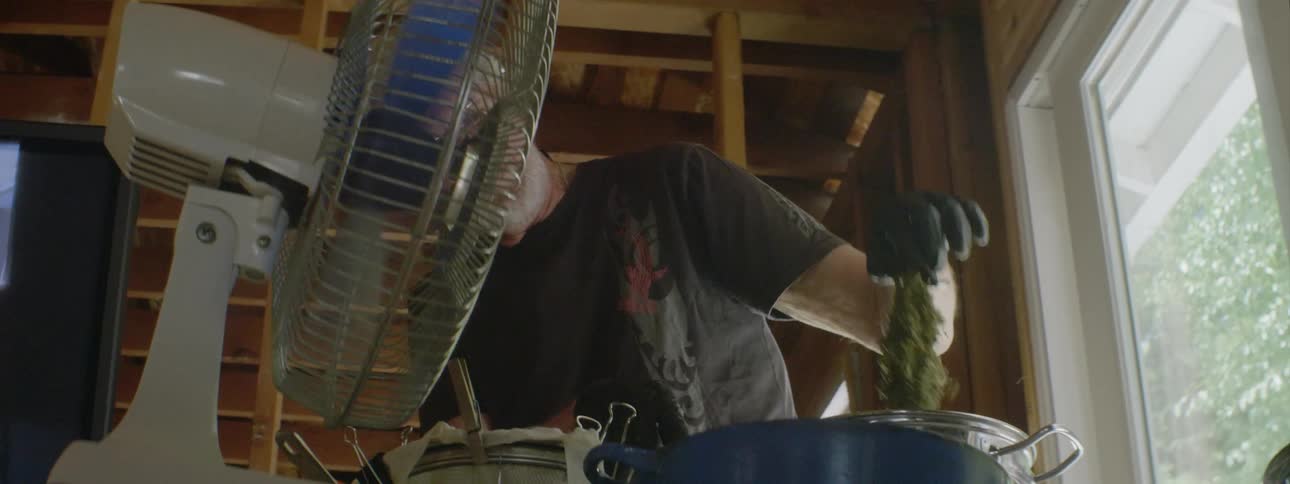 A man wearing gloves shakes off a piece of green while stirring a pot as a fan blows