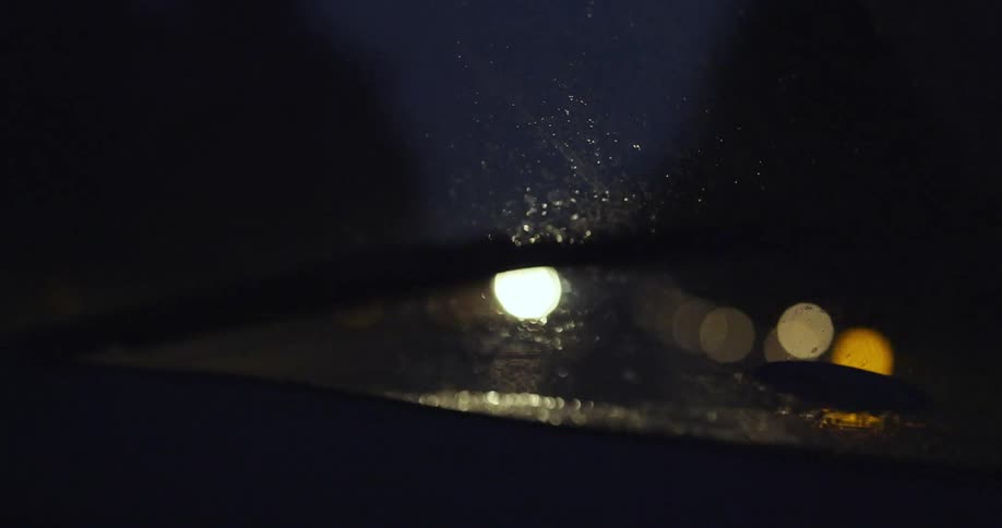 Windshield wiper works to clear rain drops from glass as vehicle passes other vehicles with glaring headlights at night