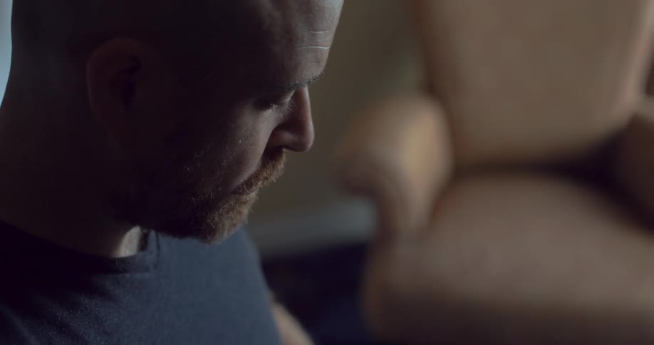 Man with a beard and a black t shirt holds and looks at the smartphone next to a brown armchair in a room