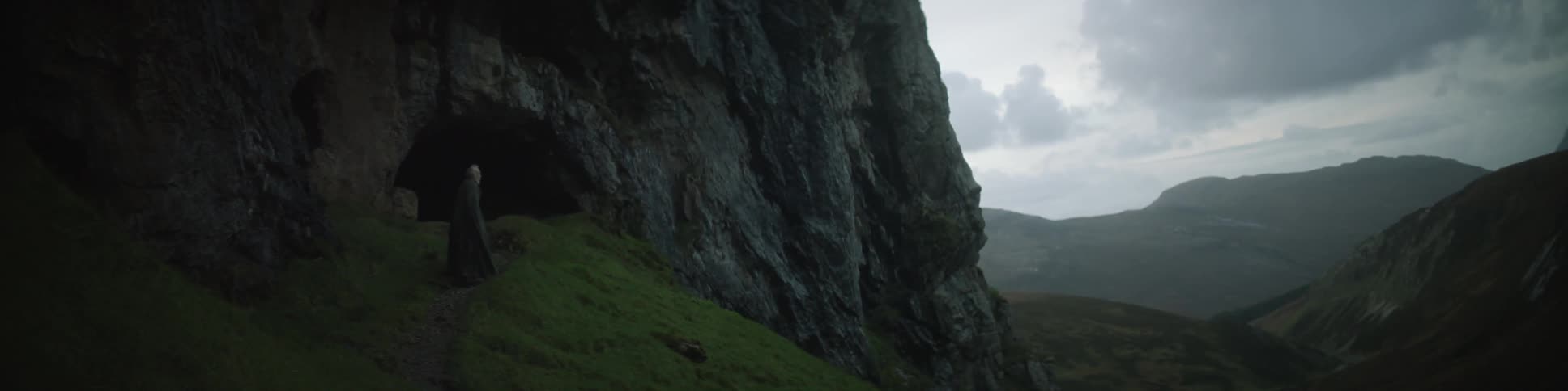 A cloaked man walking on a mountain trail that leads to a cave