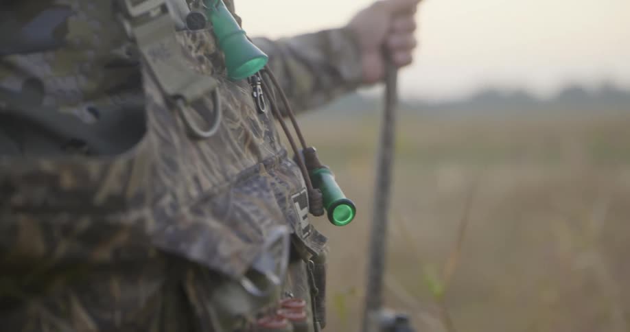A man wearing full camouflage stands in a field holding a stick