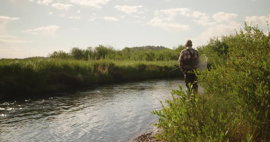 Fisherman stands on the side of a river holding a fishing pole and a fish net under his arm