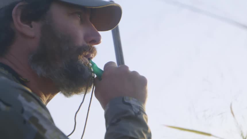 A man is sitting in a field holding a gun and uses a bird call while a dog sits nearby
