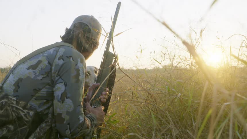 A man out hunting with his dog knees in the grass with his gun in both hands