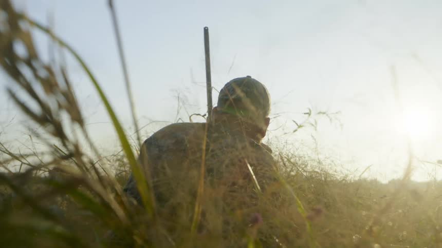 A man wearing camouflage and holding a rifle crouches down to blend into an open field