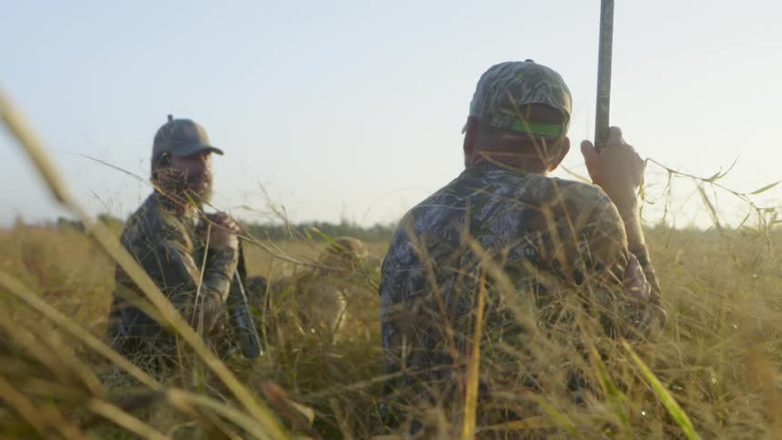 Two hunters chat as they kneel in a field holding rifles with a dog