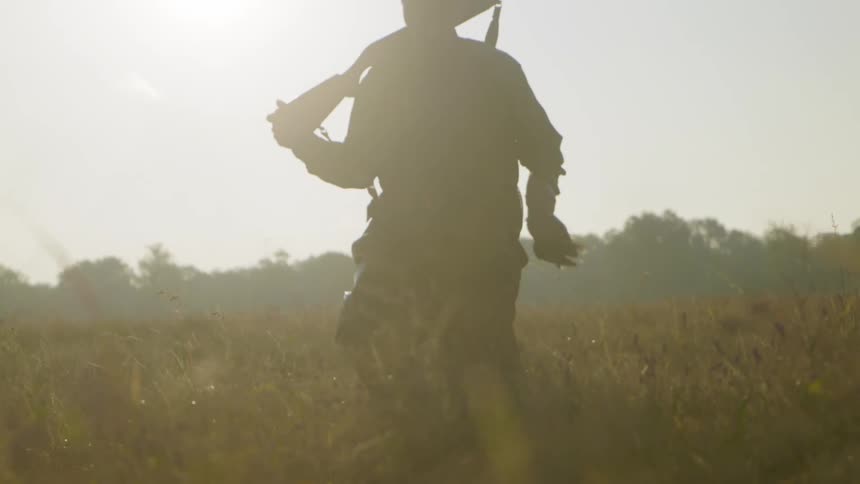 A man walks through a field with a gun on his shoulder and a dead bird in his hand