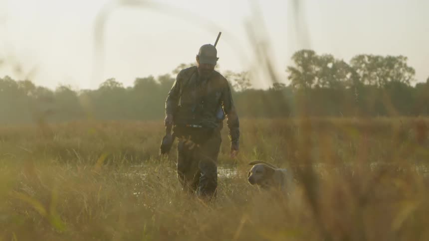 Man walking out in a wetland with his dog through water holding a dead animal carrying a hunting gun on his back