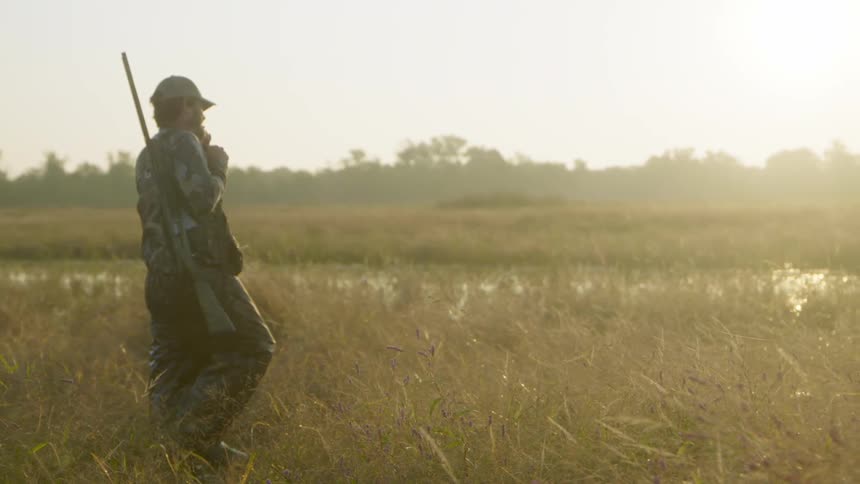 A man walks through a field and water with a gun on his shoulder and a dog next to him