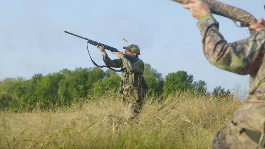 Two men sit in a field and stand up to shoot their shotguns in the air