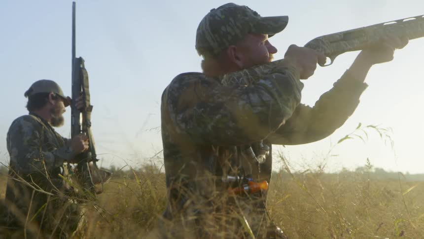 Two young white men are holding guns and shooting at something in the air and their dog goes to chase it