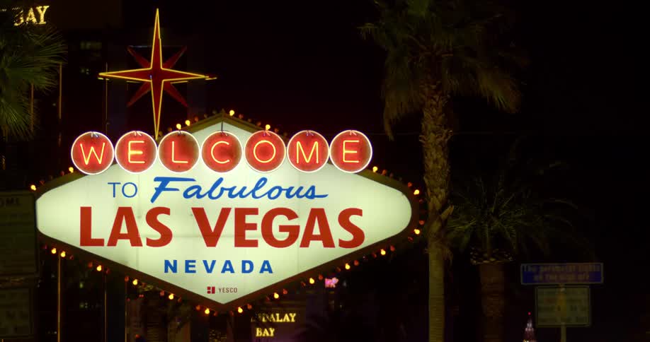 A neon red and blue welcome to las vegas sign lights up the night sky amidst palm trees blowing in the breeze