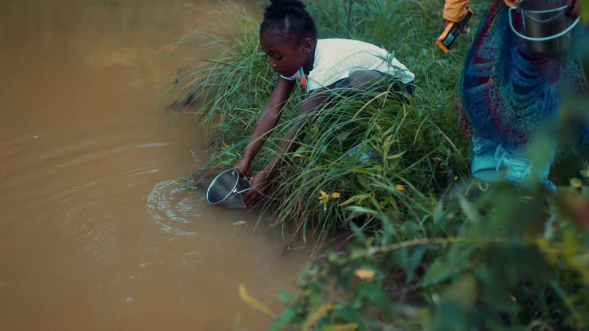 Little girl filing a bucket of water down by a murky stream in tall grass
