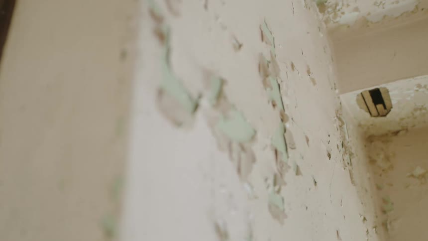 Downward view of a wall with white cracked paint above a beige floor with a large pipe