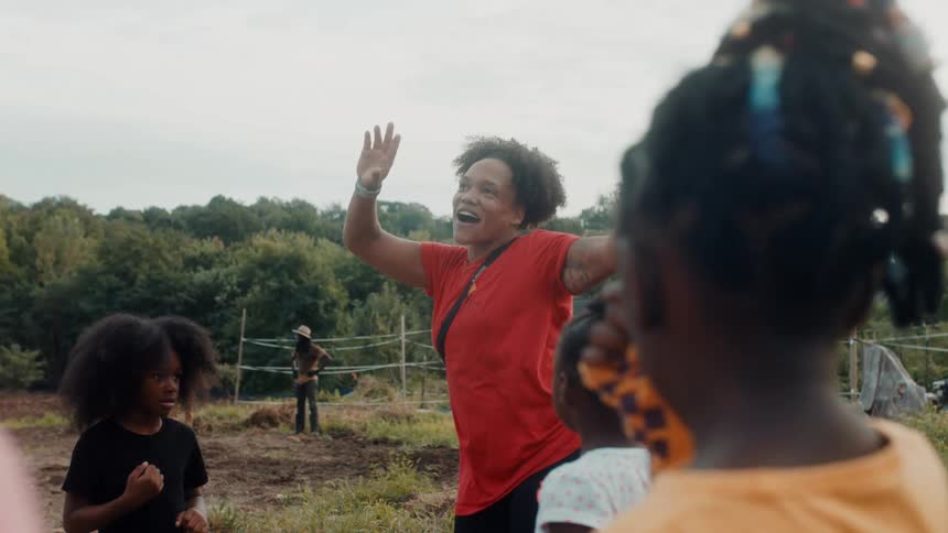 Woman and small children outside at daytime playing and dancing next to a field of bushes and grass