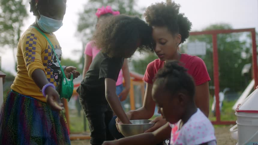A woman holds a bowl for girls to take feed and sprinkle it on the ground outside