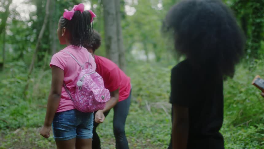 A group of children stand in line in a forest as a woman walks up and down the line and talks with them