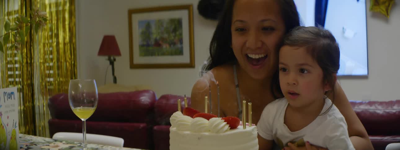 A woman and the child sitting on her lap out the lit candles on a cake in front of them