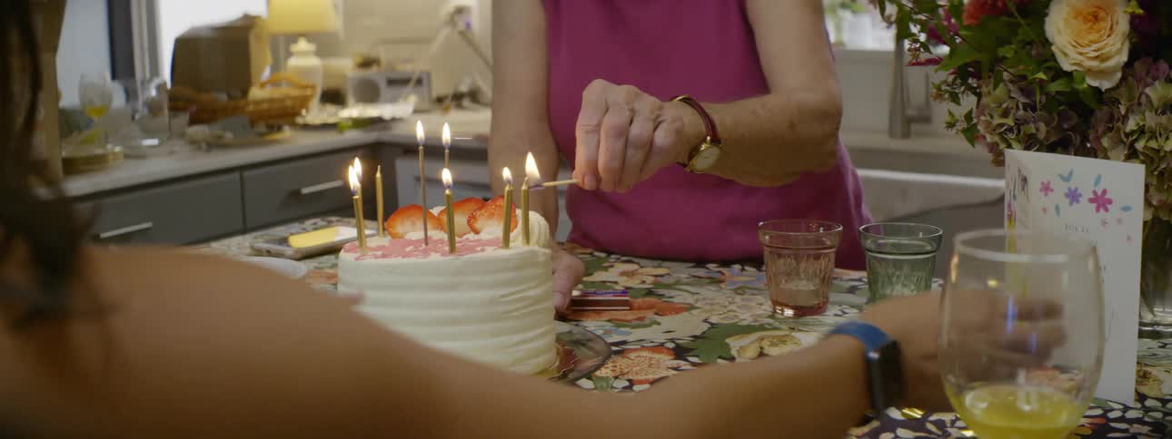 A woman lights candles on a white birthday cake sitting near a card and flowers