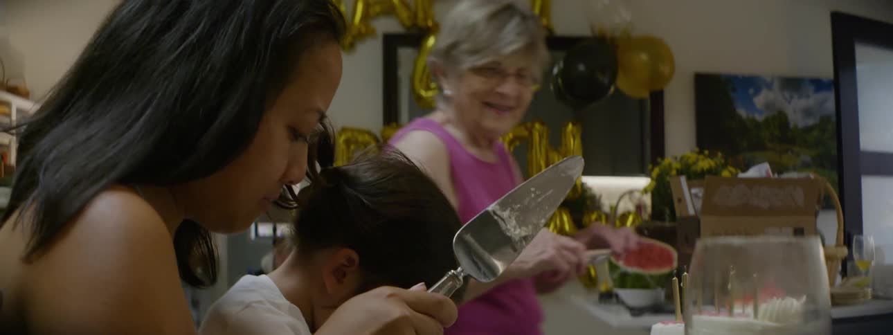 Mother and daughter sitting at table cutting a birthday cake while grandmother takes pictures