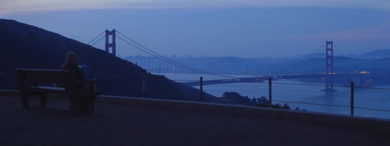 Woman sitting on a bench on an overlook drinking from a cup looking off at the golden gate bridge