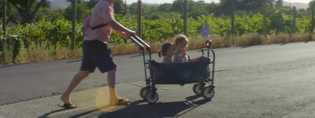 Father pushing his two daughters in a stroller towards a parked vehicle on a sunny day
