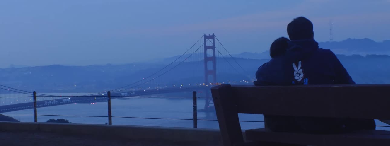 Two people cuddle on bench overlooking suspension bridge on very foggy day