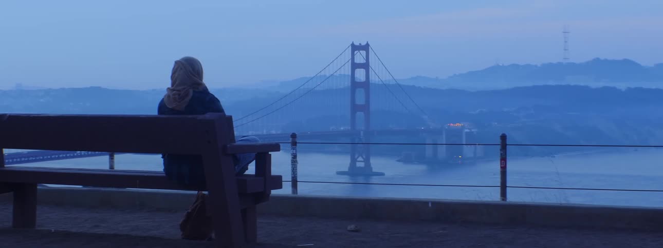 A woman sits on a bench overlooking a bridge and water at dusk with hazy conditions