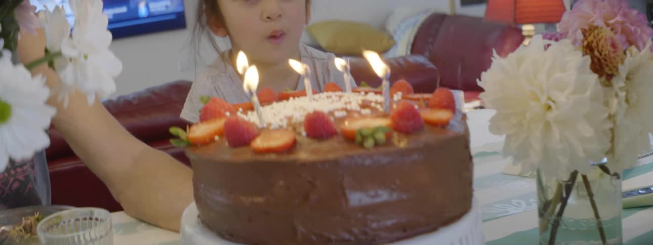 Little girl with her mother at a table blowing out candles on a chocolate birthday cake