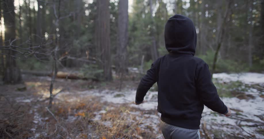 Person wearing a dark colored hoodie is walking through a forest with snow on the ground