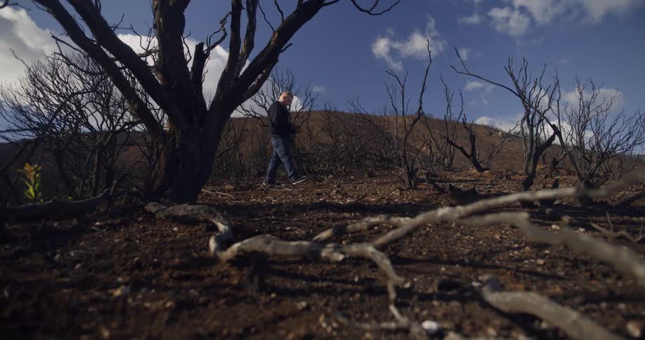 A man walks with his head down through a dirt field near hills